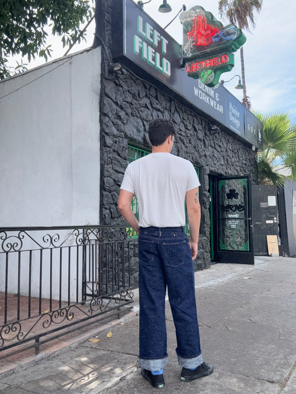 A man stands with his back to the camera wearing a white t-shirt and dark blue wide-leg jeans with rolled cuffs, positioned in front of a building with a 'LEFT FIELD WORKWEAR' sign.
