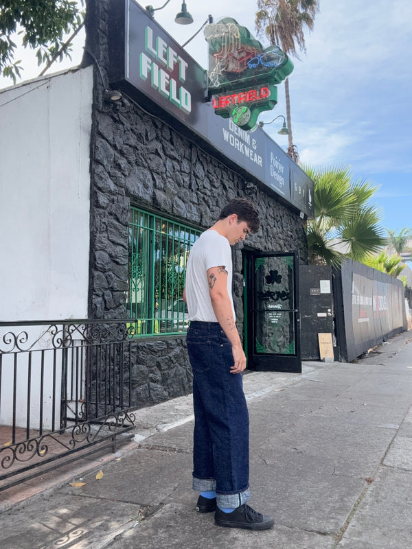 A man wearing a white t-shirt and dark blue wide-leg jeans with rolled cuffs stands on a sidewalk in front of a streetwear shop named Left Field.