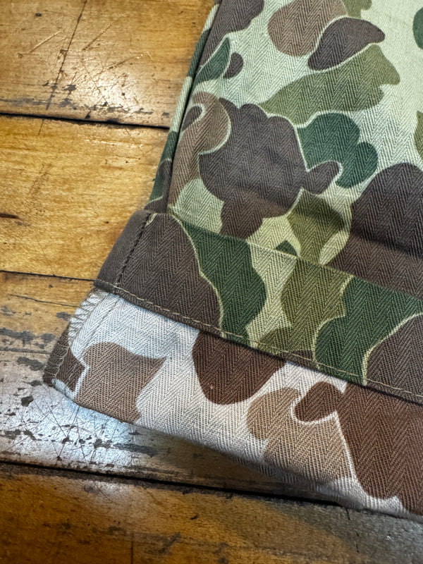Close-up of a camouflage fabric product corner folded over on a rustic wooden table. The pattern mixes olive green, brown, tan, and off-white in a herringbone-textured weave.