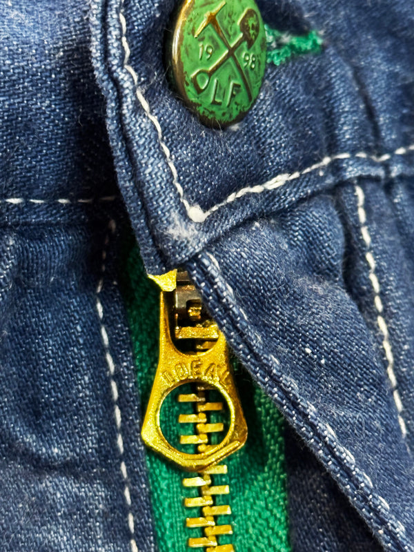 Close-up of a blue denim jacket with a gold zipper and green button.