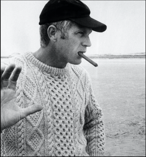 Man wearing a knitted sweater and cap, smoking a cigarette by a body of water.
