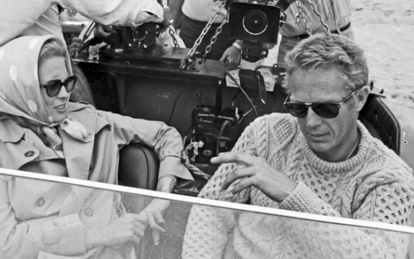 Two people sitting in a vehicle with a film crew in the background