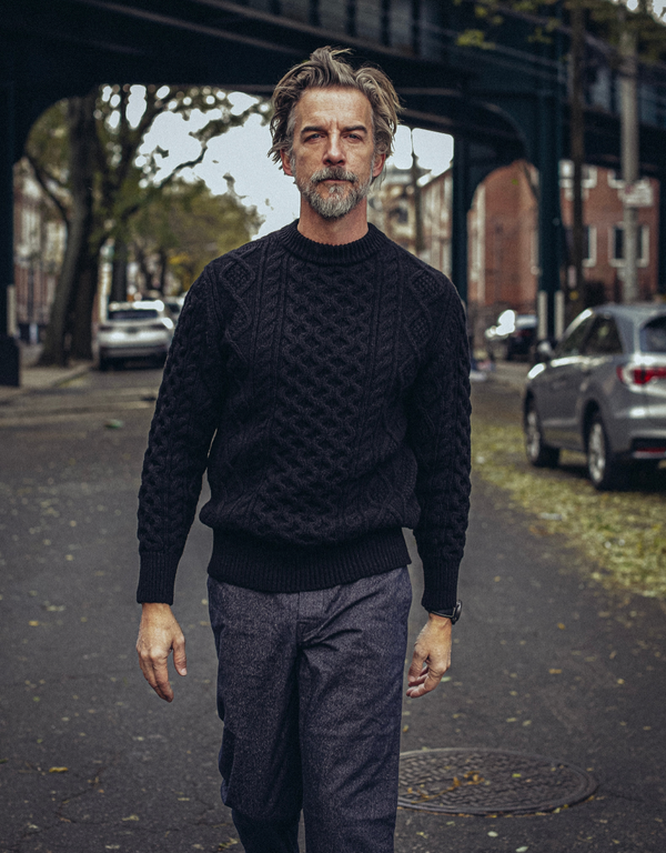 Man wearing a dark sweater and gray pants standing on a street with cars and trees in the background.