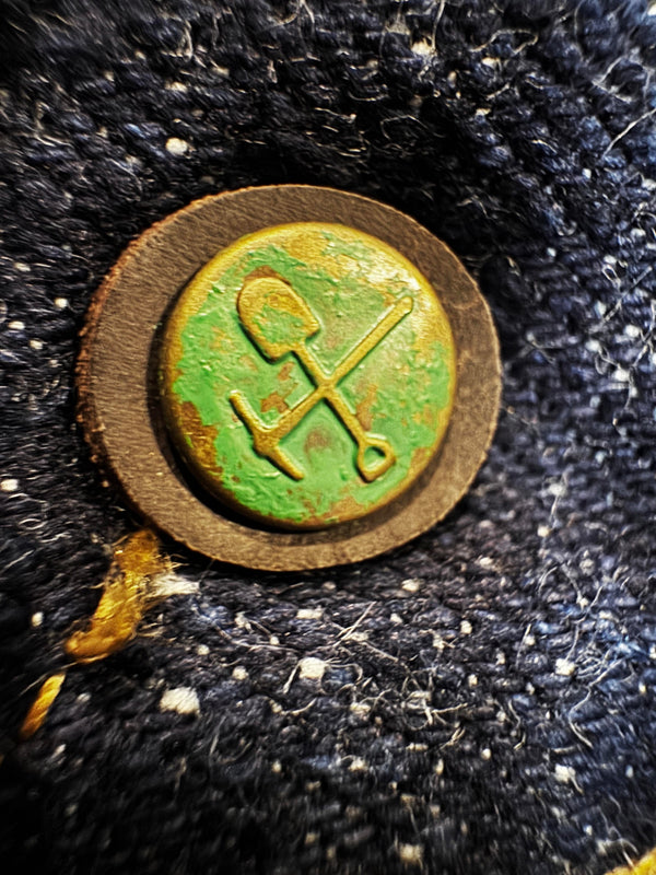A close-up of a round vintage button or badge with a green background and crossed shovel and pickaxe symbols, set against a dark textured fabric.