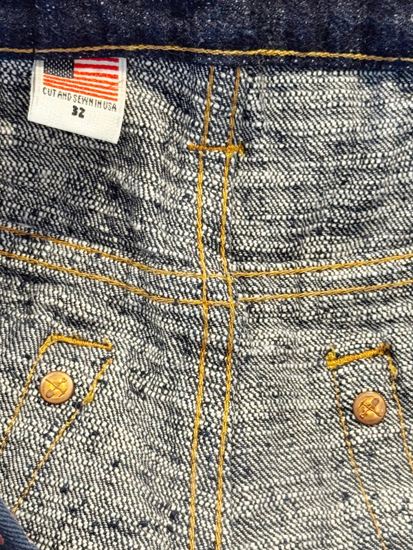 Close-up of the inside back waistband of a pair of denim jeans, showing a white size label with an American flag and 'CUT AND SEW IN USA 92' stitched in yellow contrast thread. The denim is a textured medium wash gray-blue with yellow stitching and brass buttons.