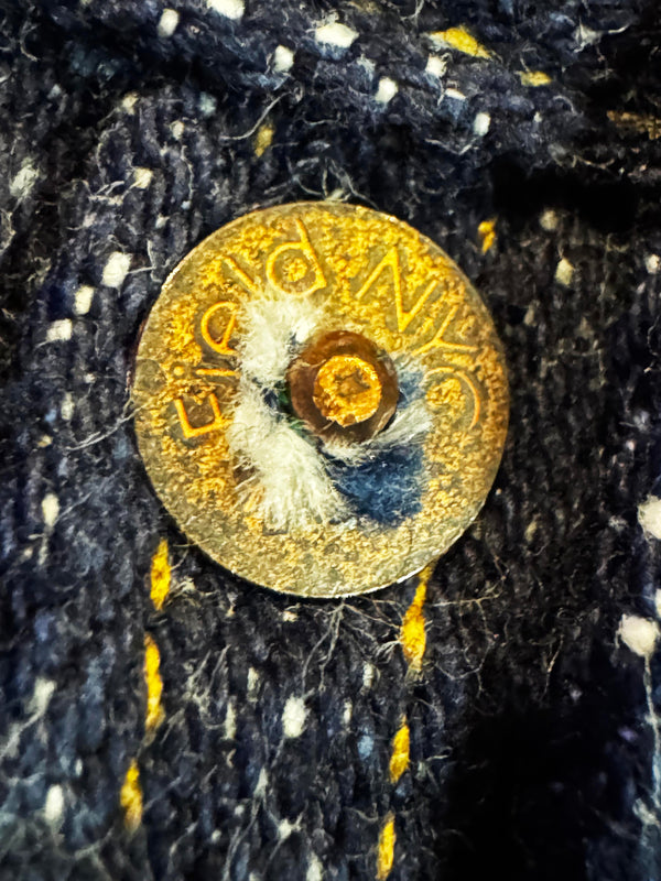 A close-up of a round gold-tone metal button with the brand name 'BIC' visible, set into dark blue and white heathered denim fabric.