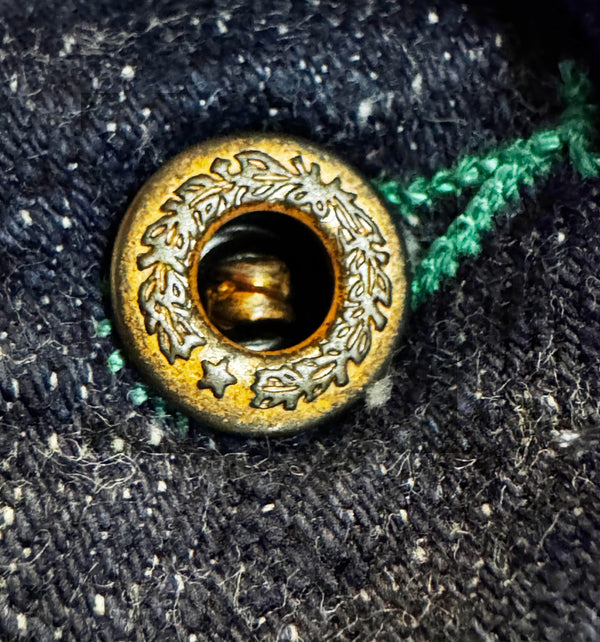 Close-up of a decorative brass button with floral engraving, set into dark textured fabric with a green thread tail visible on the right.