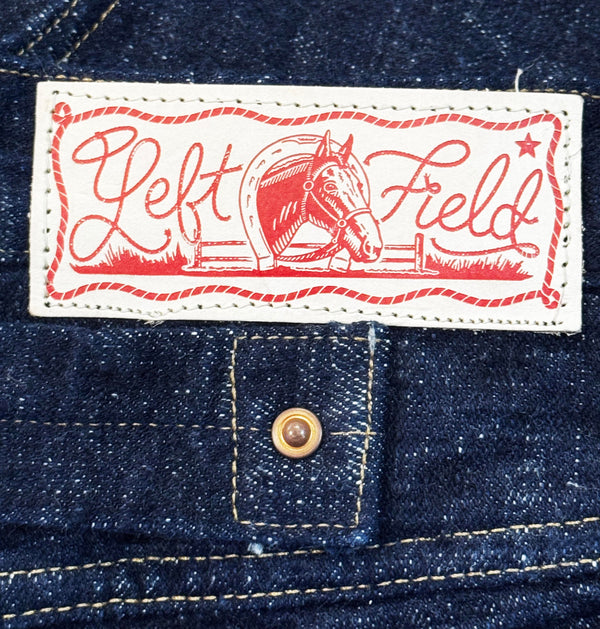Close-up of a denim garment showing a white rectangular brand patch with red script reading "Left Field" and a horse illustration, plus a metal snap button below.