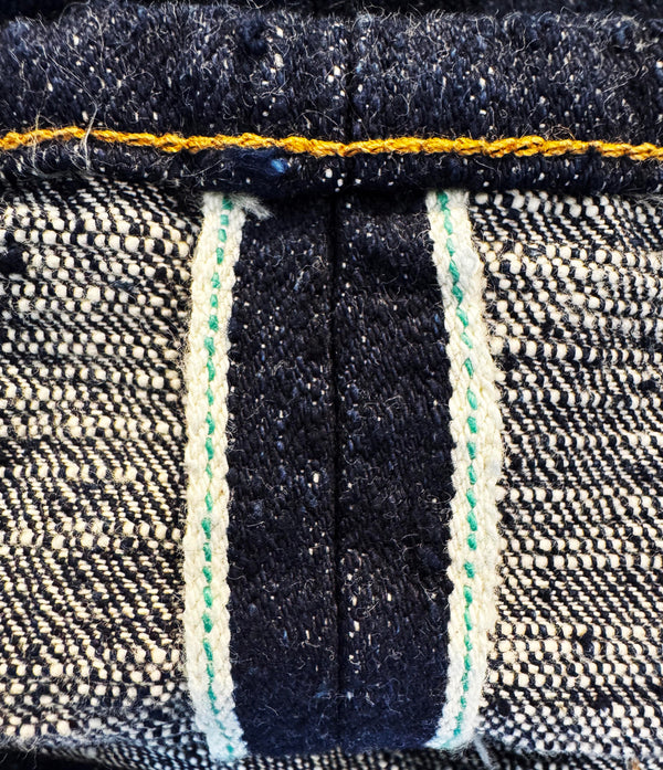 Close-up of a denim garment showing the inner waistband construction, with dark indigo denim, a yellow-orange seam, and off-white contrast stitching.