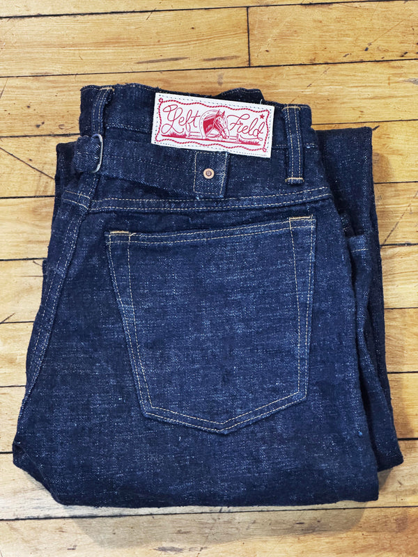 A pair of folded dark indigo denim jeans, showing the back pocket and the red and white 'Dead Felt' brand label at the waistband, laid flat on a light-colored wooden plank floor.