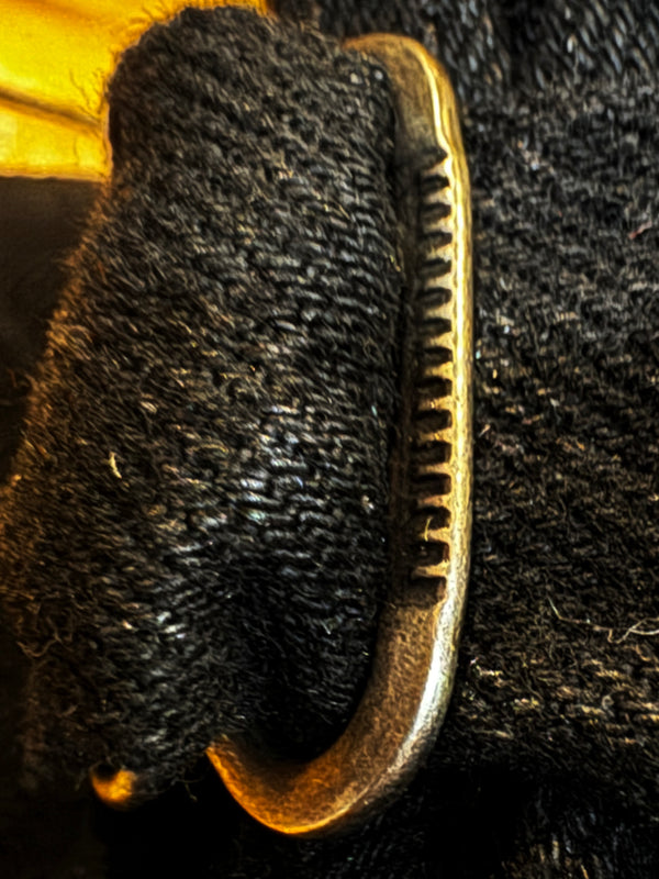 A close-up of a curved, antique brass-colored metal hook or clasp with a serrated edge, resting against a textured black fabric background.