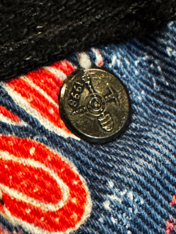 A close-up of a round metal button with a cross-shaped pin, set onto a blue denim fabric with red and white printed patterns.
