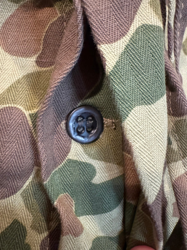 Close-up of a camouflage-patterned fabric garment with a dark button, showing shades of green, brown, tan, and beige.