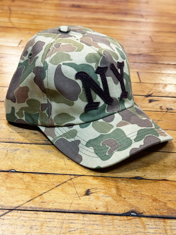 A baseball cap with a green, brown, and beige camouflage pattern, featuring a large black embroidered 'NY' logo on the front, displayed on a wooden floor.