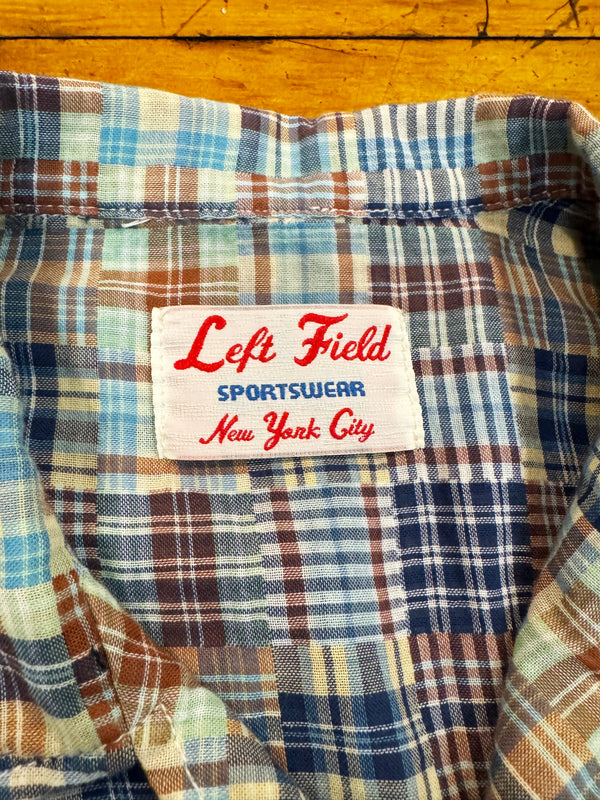 Close-up of a plaid shirt collar with a visible 'Left Field Sportswear New York City' brand label, showing a blue, brown, beige, and white plaid pattern against a yellow wooden background.