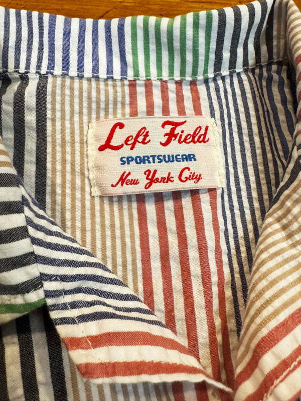 Close-up of a striped shirt collar with a visible brand label reading Left Field Sportswear New York City. The fabric has vertical stripes in cream, navy, red, green, and tan.