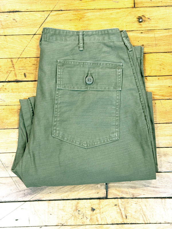 A pair of olive green pants folded flat on a light-colored wooden plank surface, showing a buttoned back pocket.
