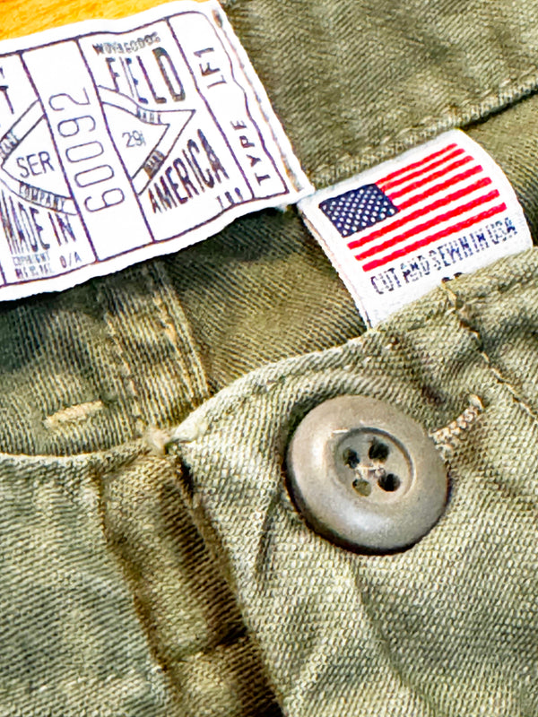 Close-up of olive green denim fabric showing a white woven label with black text, a small American flag patch, and a gray metal button.