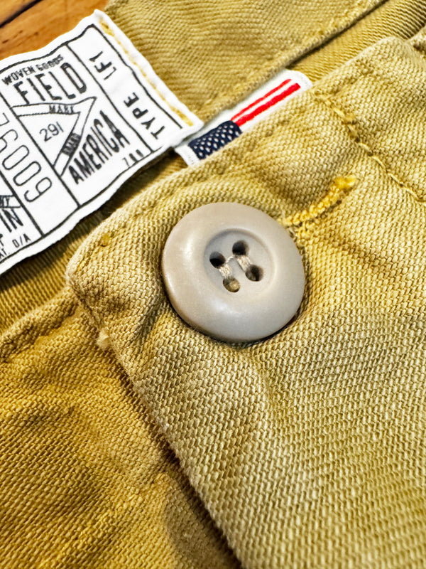 Close-up of khaki woven fabric with a light gray button, a white sewn label reading Western Crowns Field Type I, and a small American flag tag.