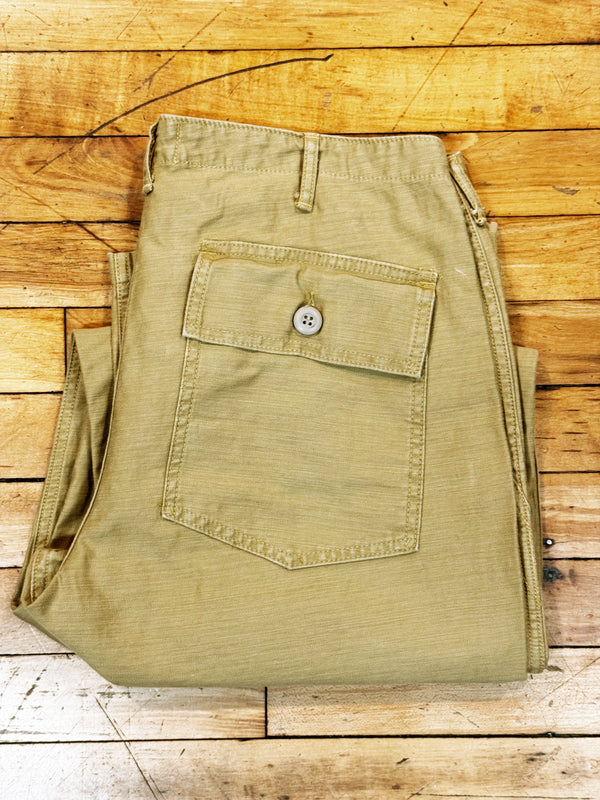 A pair of folded khaki pants with a buttoned front flap pocket, displayed on a rustic wooden plank background.