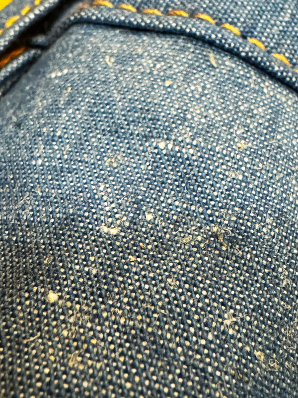 Close-up of dark blue denim fabric with visible yellow contrast stitching and small lint or fiber particles on the surface.